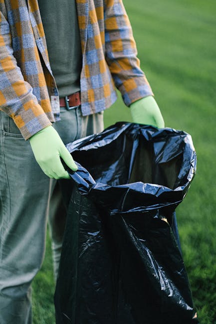 A person standing outdoors on a grassy area, wearing a plaid shirt over a green t-shirt, with light grey trousers secured by a brown belt. They are wearing light green gloves and holding a large black
